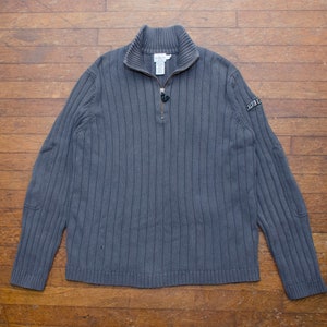 Peut inclure: Un pull en tricot côtelé gris anthracite avec une fermeture éclair quart de zip et un col montant. Le pull présente un petit écusson logo sur la manche gauche. Le vêtement est présenté sur une surface en bois.