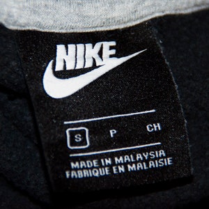 May include: Black and white Nike logo tag with the text "NIKE" in white. The tag also includes the size "S", "P", and "CH" in white. The tag reads "Made in Malaysia" and "Fabrique en Malaisie" in white.