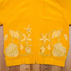 以下が含まれることがあります： 鮮やかな黄色のジャケット。クリーム色とゴールドの色合いで貝殻とヒトデの刺繍が施されています。ロープのようなアクセントと小さな金のビーズがデザインに取り入れられています。夏の装いに最適です。