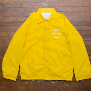 May include: A bright yellow coach jacket with a button-up front and a collar. The jacket features the text "ROB'S BODY SHOP" and "Athens, Ohio" in white lettering. The jacket is made of a lightweight material.