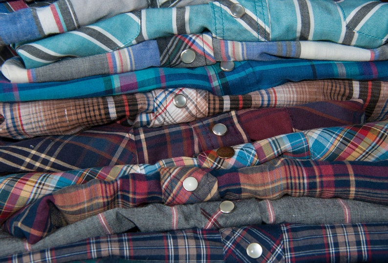 May include: A stack of plaid shirts in various colours, including blue, red, and brown. The shirts are folded neatly and have metal buttons.