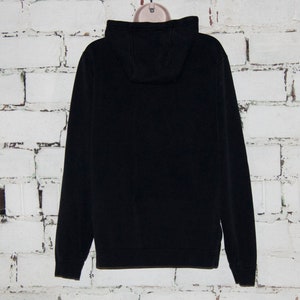 May include: A black hooded sweatshirt with a zipper closure. The sweatshirt is hanging on a white brick wall.