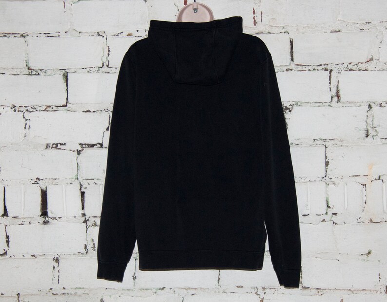 May include: A black hooded sweatshirt with a zipper closure. The sweatshirt is hanging on a white brick wall.