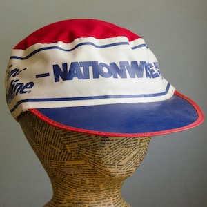 May include: A vintage cycling cap with a red top, white midsection, and blue brim. The white section features the word "NATIONWIDE" in blue block letters. The brim is trimmed with red. The cap is displayed on a mannequin head.