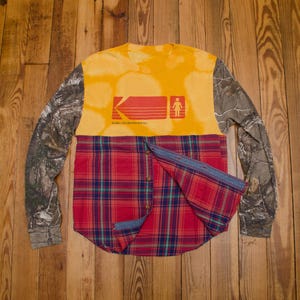 Puede incluir: Una camisa de manga larga con mangas de camuflaje, una sección superior amarilla en el pecho con un logotipo gráfico rojo de Girl Skateboards y una sección inferior de cuadros rojos, azules y verdes con una tapeta de botones frontal.