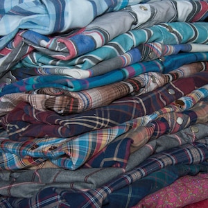 May include: A stack of plaid and striped shirts in various shades of blue, red, and brown. The shirts are folded and stacked on top of each other, creating a layered effect.