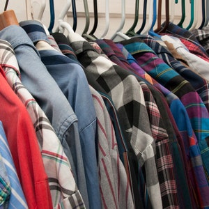 May include: A collection of plaid shirts in various colours and patterns hanging on a rack. The shirts are all long-sleeved and have a button-down collar.