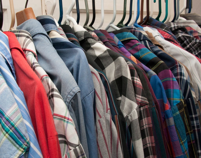 May include: A collection of plaid shirts in various colours and patterns hanging on a rack. The shirts are all long-sleeved and have a button-down collar.