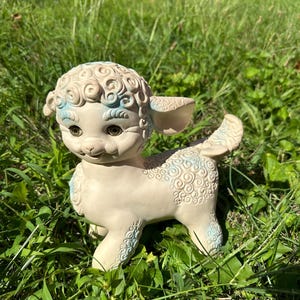 Peut inclure: Un agneau en plastique blanc et bleu avec des cheveux bouclés et un visage souriant. L'agneau est debout dans un champ herbeux.