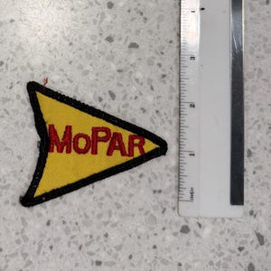 Puede incluir: Parche bordado amarillo y negro con la palabra "MoPAR" en letras rojas. El parche tiene forma triangular. Una regla está al lado del parche.