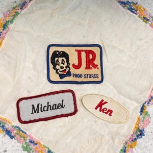 Könnte beinhalten: Ein weißes Vintage-Taschentuch mit farbenfrohem Häkelrand. Es zeigt drei gestickte Aufnäher: ein "JR FOOD STORES"-Logo und Namensschilder mit den Aufschriften "Michael" und "Ken".