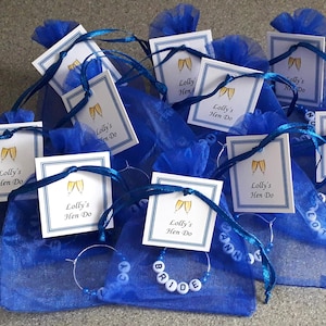 May include: A collection of blue organza drawstring bags, each containing a circular metal wine glass charm with white lettered beads. Attached to each bag is a square white tag with a blue border, featuring two gold champagne flutes and the text Lolly's Hen Do.