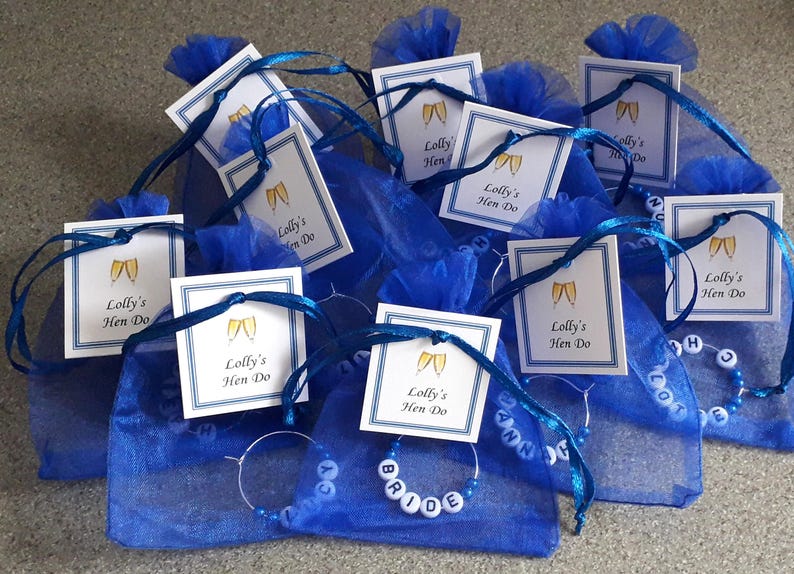 May include: A collection of blue organza drawstring bags, each containing a circular metal wine glass charm with white lettered beads. Attached to each bag is a square white tag with a blue border, featuring two gold champagne flutes and the text Lolly's Hen Do.