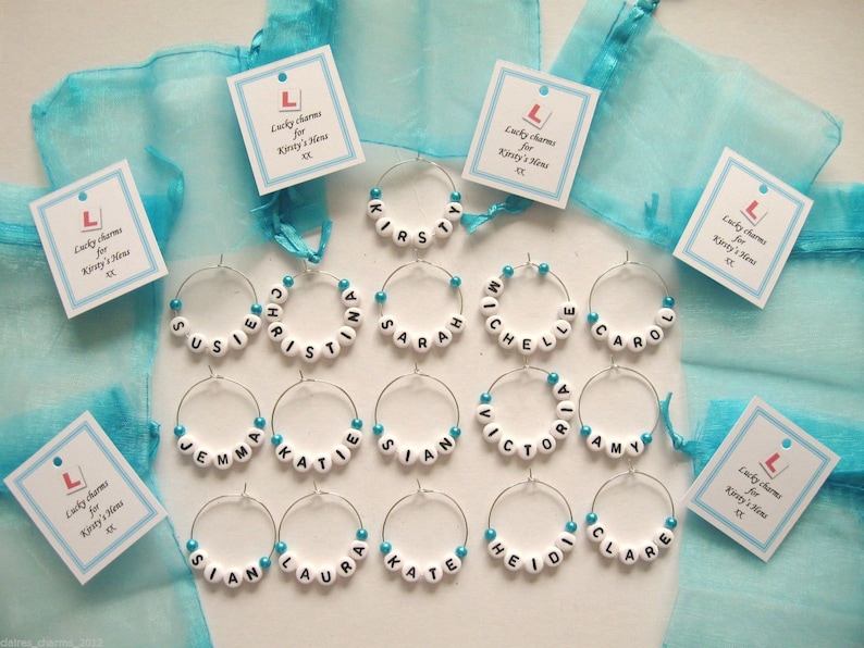 May include:  wine glass charms with white beads spelling out different names. The charms are displayed on a turquoise background with small cards that say "Lucky charms for Kirsty's New" with a red L.