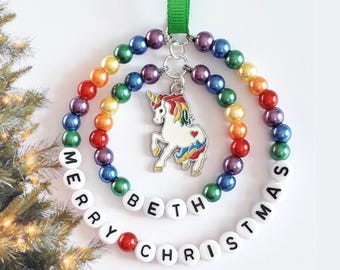 May include: A double-ring Christmas ornament featuring colorful metallic beads and white letter beads spelling MERRY CHRISTMAS and BETH. A silver-toned unicorn charm with a rainbow mane and tail hangs from the center, suspended by a green ribbon.