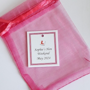 May include: A sheer pink organza drawstring pouch featuring a white rectangular tag with a red L-plate symbol and the printed text: Sophie's Hen Weekend May 2024. The tag has a small hole at the top and a thin red border around the edge.