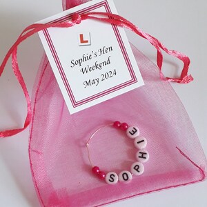 May include: A pink organza drawstring bag with a white tag featuring a red L-plate icon and the text Sophie's Hen Weekend May 2024. Inside the bag is a circular metal wine glass charm featuring pink beads and white letter beads spelling Sophie.