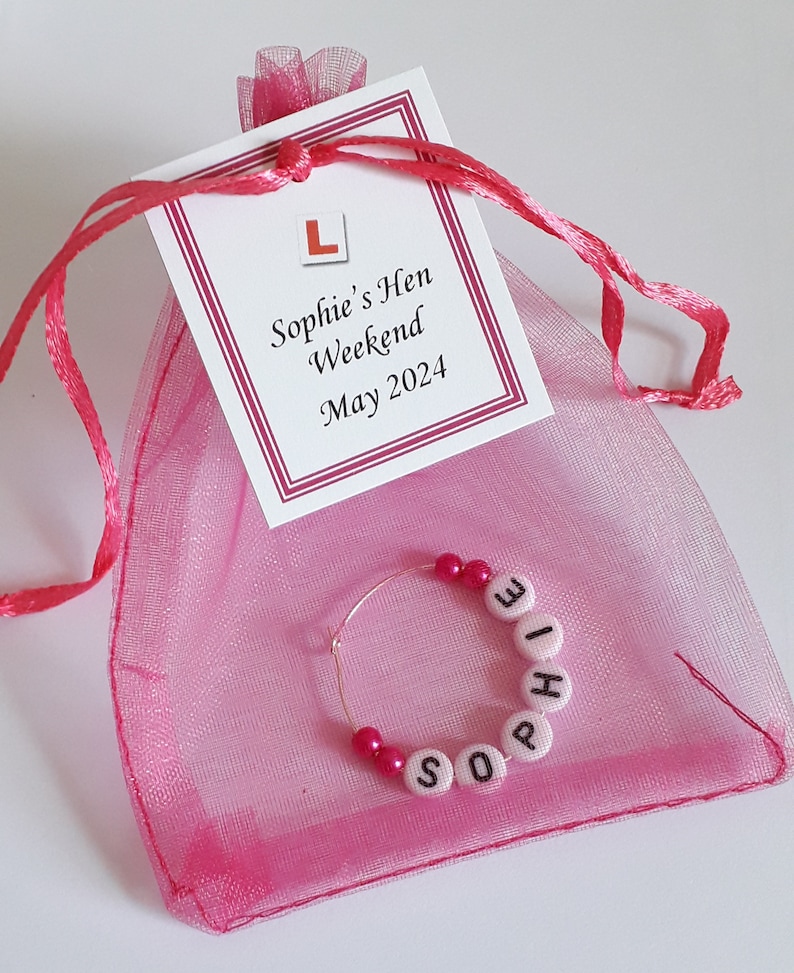 May include: A pink organza drawstring bag with a white tag featuring a red L-plate icon and the text Sophie's Hen Weekend May 2024. Inside the bag is a circular metal wine glass charm featuring pink beads and white letter beads spelling Sophie.