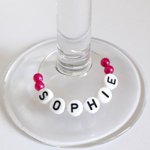 May include: A circular wine glass charm featuring a silver wire hoop decorated with white round beads displaying the black printed name SOPHIE and two pairs of small, shiny, hot pink spherical beads positioned at the ends of the wire.