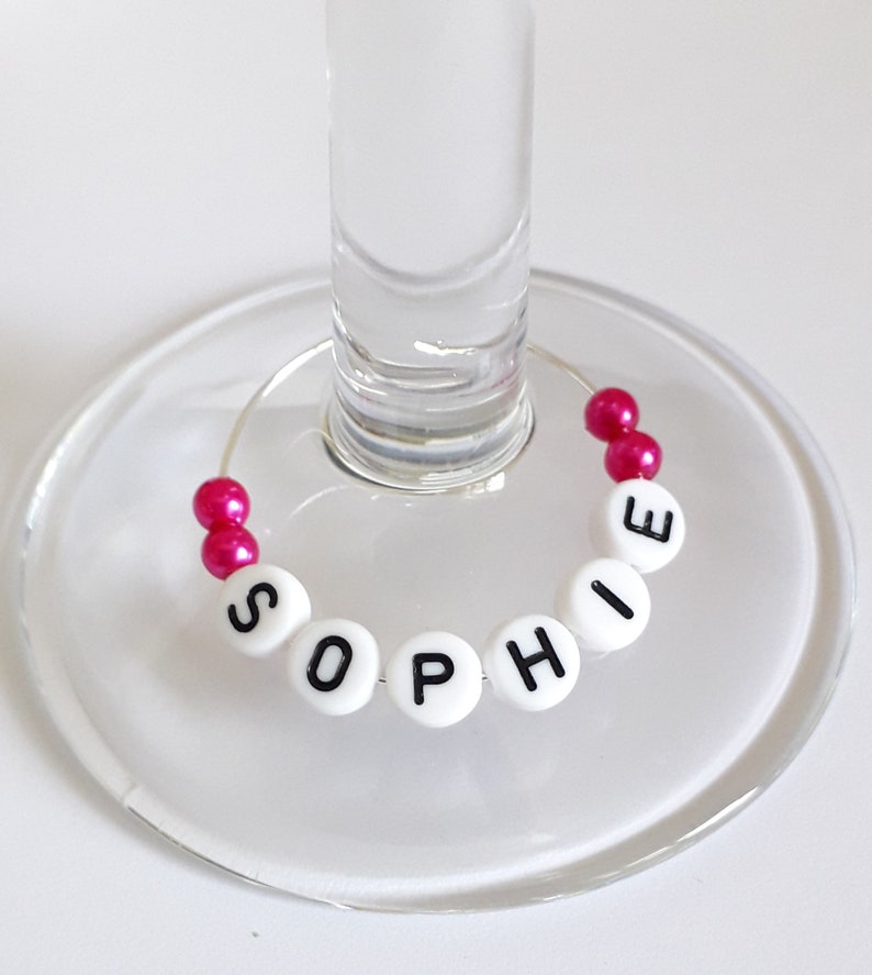 May include: A circular wine glass charm featuring a silver wire hoop decorated with white round beads displaying the black printed name SOPHIE and two pairs of small, shiny, hot pink spherical beads positioned at the ends of the wire.