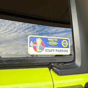 May include: A blue and yellow Vault-Tec Industries staff parking sign featuring a cartoon character giving a thumbs up. The sign reads "Vault-Tec Industries" and "Staff Parking" with a logo and the year 2025.
