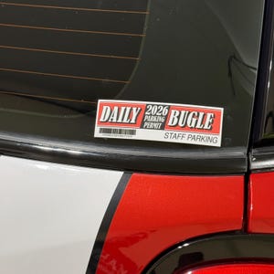 Puede incluir: Una pegatina rectangular en la ventana de un coche con las palabras "DAILY BUGLE" en rojo y blanco. La pegatina también dice "2026 PARKING PERMIT STAFF PARKING" y tiene un código de barras. El coche es blanco y rojo.