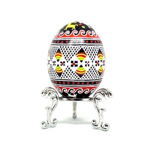 May include: A hand-painted Easter egg with a geometric design in red, yellow, black, and white on a black background. The egg is on a silver stand.