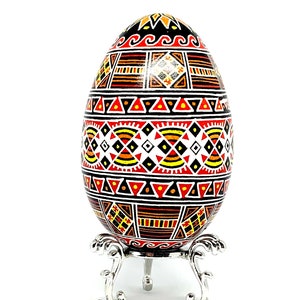 May include: A hand-painted Easter egg with a geometric pattern in black, red, orange, and yellow on a white background. The egg is displayed on a silver stand.