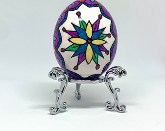 May include: A white egg decorated with a colorful floral design in shades of purple, blue, yellow, and green. The egg is displayed on a silver stand.