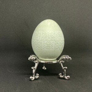May include: A white egg with a decorative pattern is displayed on a silver egg stand. The egg has a raised, intricate design.