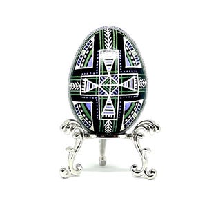 May include: A hand-painted Easter egg with a geometric design in shades of green, purple and white on a black background. The egg is displayed on a silver stand with three legs.