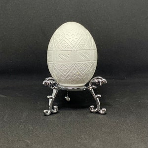 May include: A white ceramic egg with an intricate carved design sits on a silver stand with three legs.