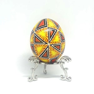 May include: A yellow, orange, and black decorated Easter egg on a silver egg stand. The egg has a geometric pattern of triangles and sunbursts.