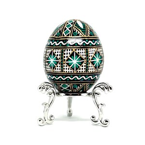 May include: A green and gold decorated Easter egg on a silver stand. The egg has a geometric pattern with white accents.