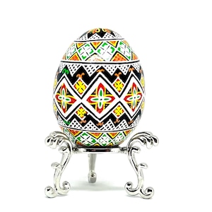 May include: A hand-painted Easter egg with a geometric pattern in black, white, red, yellow, and green colours. The egg is displayed on a silver stand.