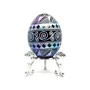 May include: A black and white patterned Easter egg with a iridescent purple and blue sheen, displayed on a silver stand.