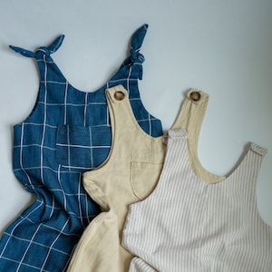 May include: Three children's jumpsuits in different colors and patterns. One is blue with a white grid pattern, another is beige, and the third is beige with vertical stripes. Each has a pocket and button details.