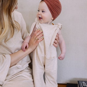 May include: A baby wearing a beige jumpsuit with front pockets and a rust-colored turban. The baby is being held by a person wearing a matching beige outfit. The baby is smiling and looking to the side.