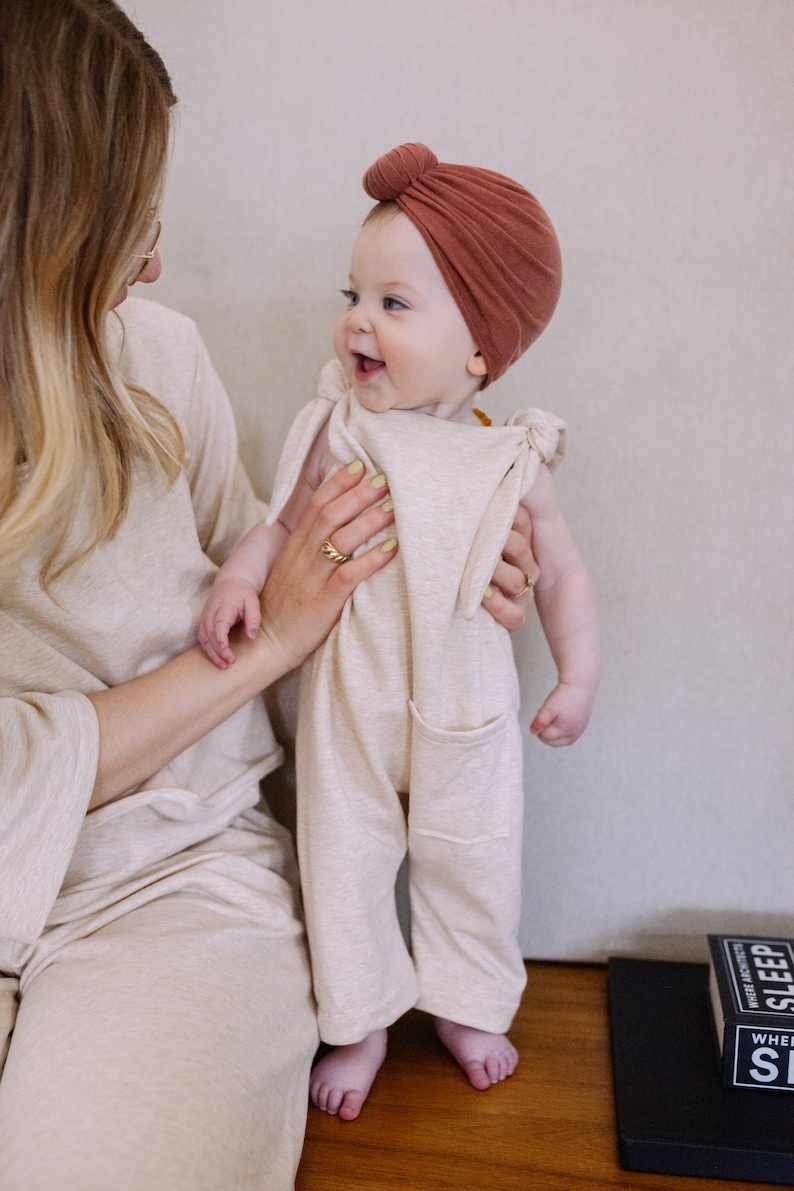 May include: A baby wearing a beige jumpsuit with front pockets and a rust-colored turban. The baby is being held by a person wearing a matching beige outfit. The baby is smiling and looking to the side.