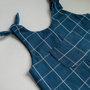 May include: Close-up of a blue and white plaid top with tie straps. The top features a small pocket and a grid pattern. The straps are tied in bows at the shoulders. The fabric appears to be a soft cotton or linen.
