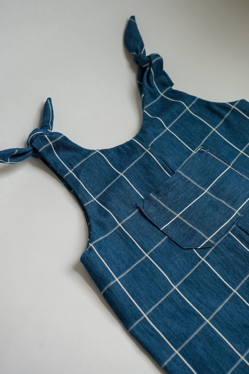 May include: Close-up of a blue and white plaid top with tie straps. The top features a small pocket and a grid pattern. The straps are tied in bows at the shoulders. The fabric appears to be a soft cotton or linen.