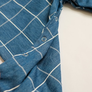 May include: Close-up of a blue denim fabric with a white grid pattern. The fabric is folded, revealing a row of silver snap closures along the edge. The overall texture appears soft and slightly worn.