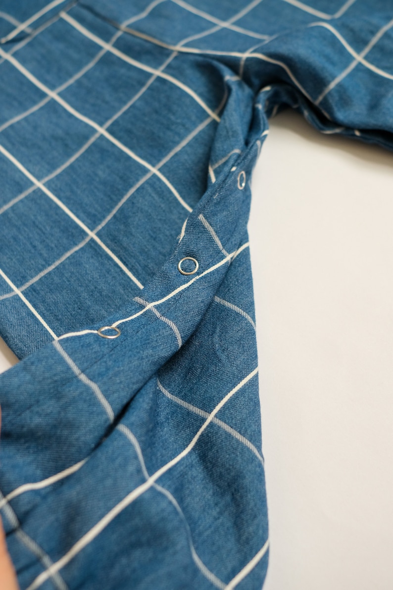 May include: Close-up of a blue denim fabric with a white grid pattern. The fabric is folded, revealing a row of silver snap closures along the edge. The overall texture appears soft and slightly worn.