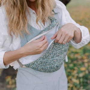 May include: A woman is holding a blue and white floral fabric bag with a zipper. The bag is worn as a crossbody bag. The woman is wearing a white shirt and blue overalls.