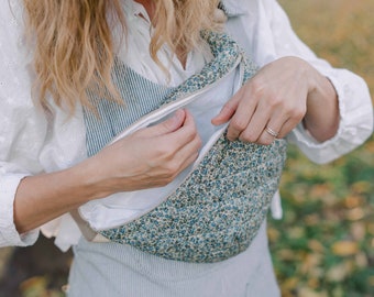 May include: A woman is holding a blue and white floral fabric bag with a zipper. The bag is worn as a crossbody bag. The woman is wearing a white shirt and blue overalls.