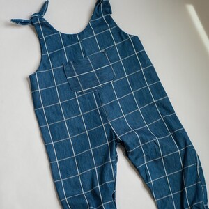 May include: Blue denim jumpsuit with a white grid pattern. The straps are tied at the shoulders, and there is a pocket on the chest. The jumpsuit has a relaxed fit and is suitable for children's wear.