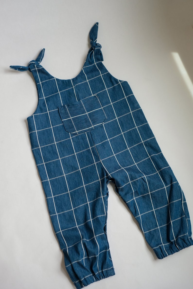 May include: Blue denim jumpsuit with a white grid pattern. The straps are tied at the shoulders, and there is a pocket on the chest. The jumpsuit has a relaxed fit and is suitable for children's wear.