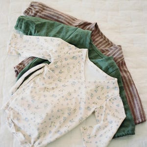 Könnte beinhalten: Eine Auswahl an Babykleidung in verschiedenen Farben und Mustern. Das oberste Kleidungsstück ist ein cremefarbener Strampler mit blauem Blumenmuster. Weitere Artikel sind ein grünes Hemd und ein braunes gestreiftes Oberteil. Alle Artikel sind aus weichem Stoff.