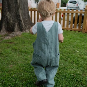 May include: A child wearing a teal linen jumpsuit with two front pockets and a white t-shirt underneath. The child is walking on green grass near a wooden fence and a large tree.