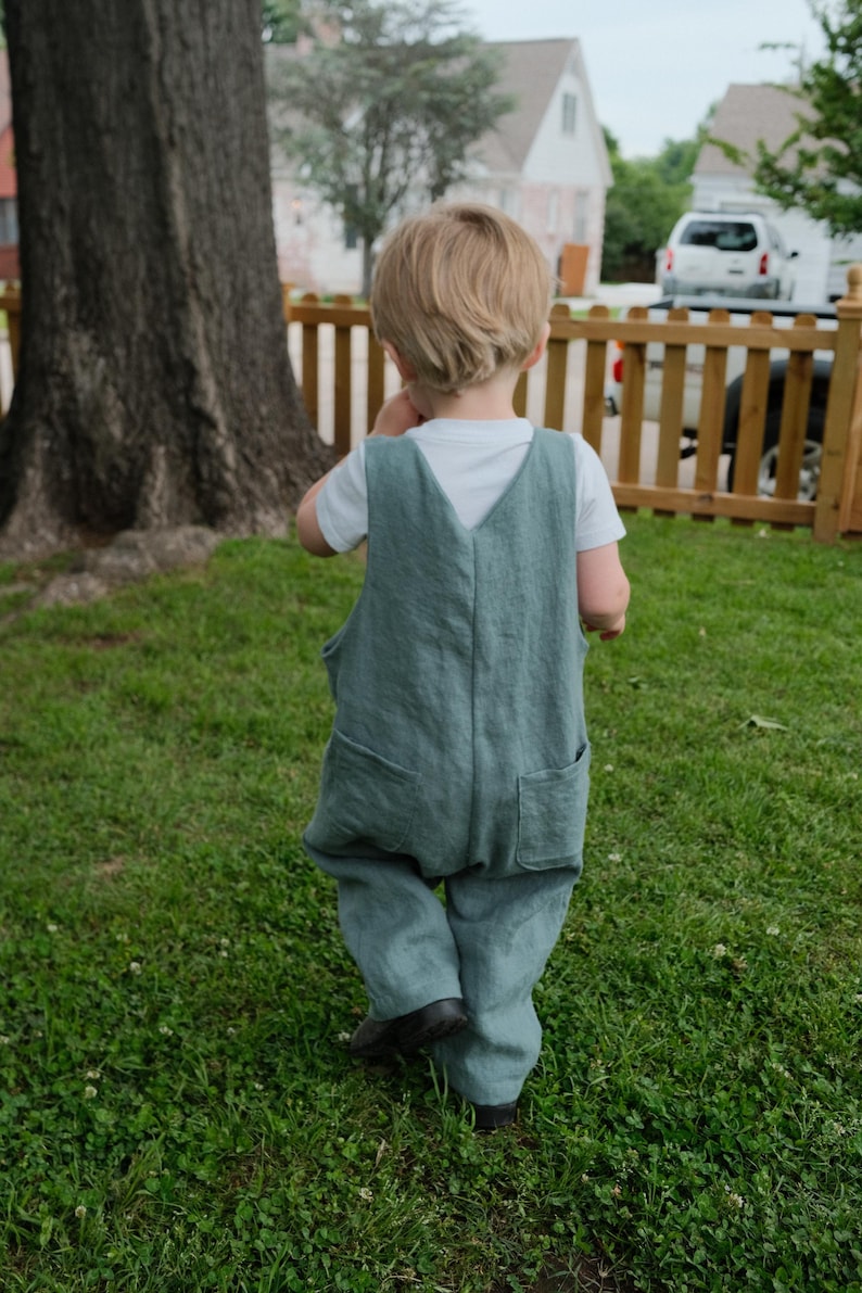 May include: A child wearing a teal linen jumpsuit with two front pockets and a white t-shirt underneath. The child is walking on green grass near a wooden fence and a large tree.